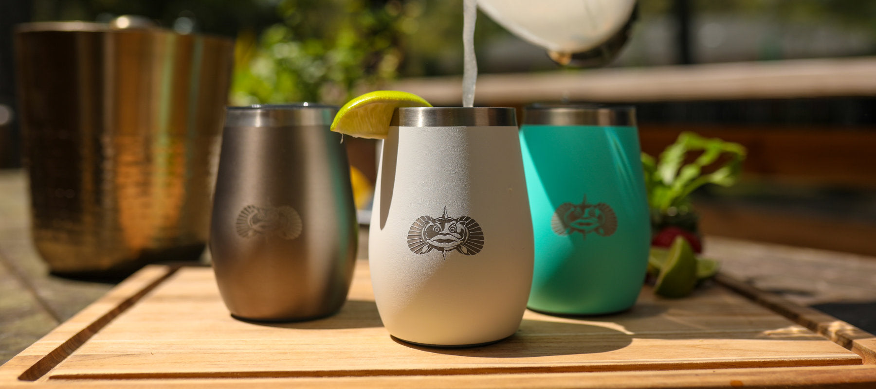 Three stainless steel tumblers on a wooden surface with a blurred outdoor background